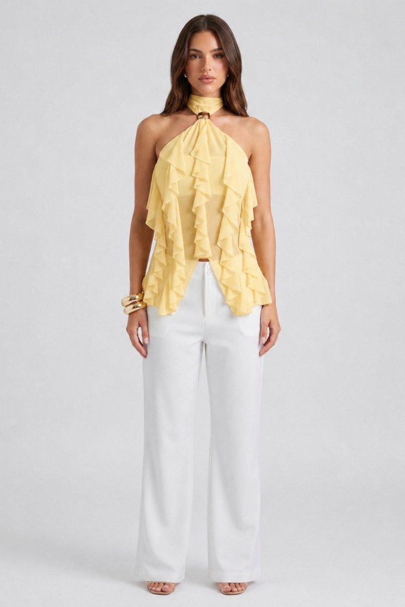 A woman modeling a pale yellow ruffled halter top with a high neck, paired with white wide-leg pants, against a light gray studio background.