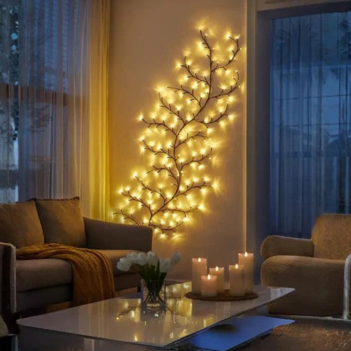 A decorative wall-mounted tree branch light fixture covered in warm glowing fairy lights, displayed in a cozy living room setting with sofas, a coffee table, and candles.