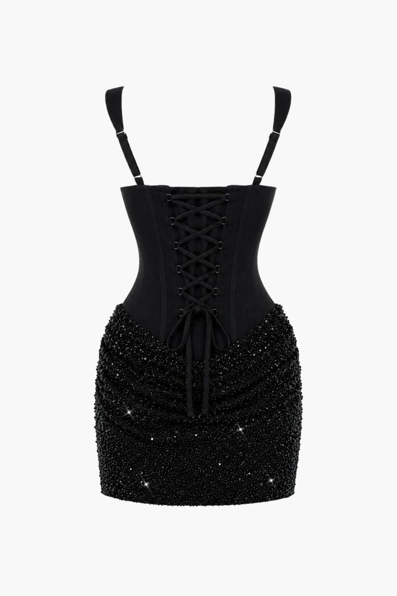 A black corset-style mini dress with thin shoulder straps, a lace-up front, and a heavily embellished beaded skirt, shown against a plain light gray background.