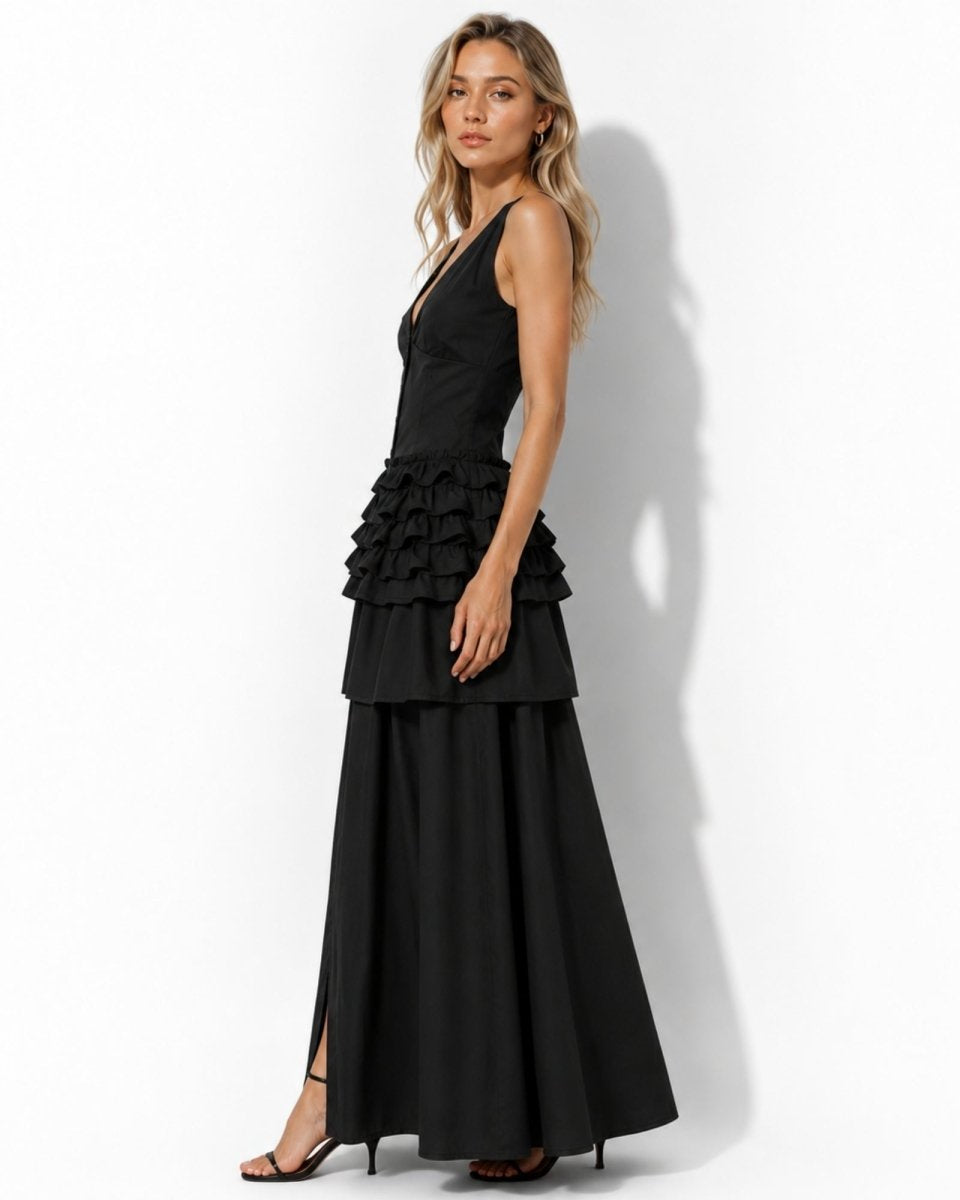 A woman modeling a black sleeveless maxi dress with a tiered ruffle waist and a long flowing skirt, standing against a plain white studio wall.