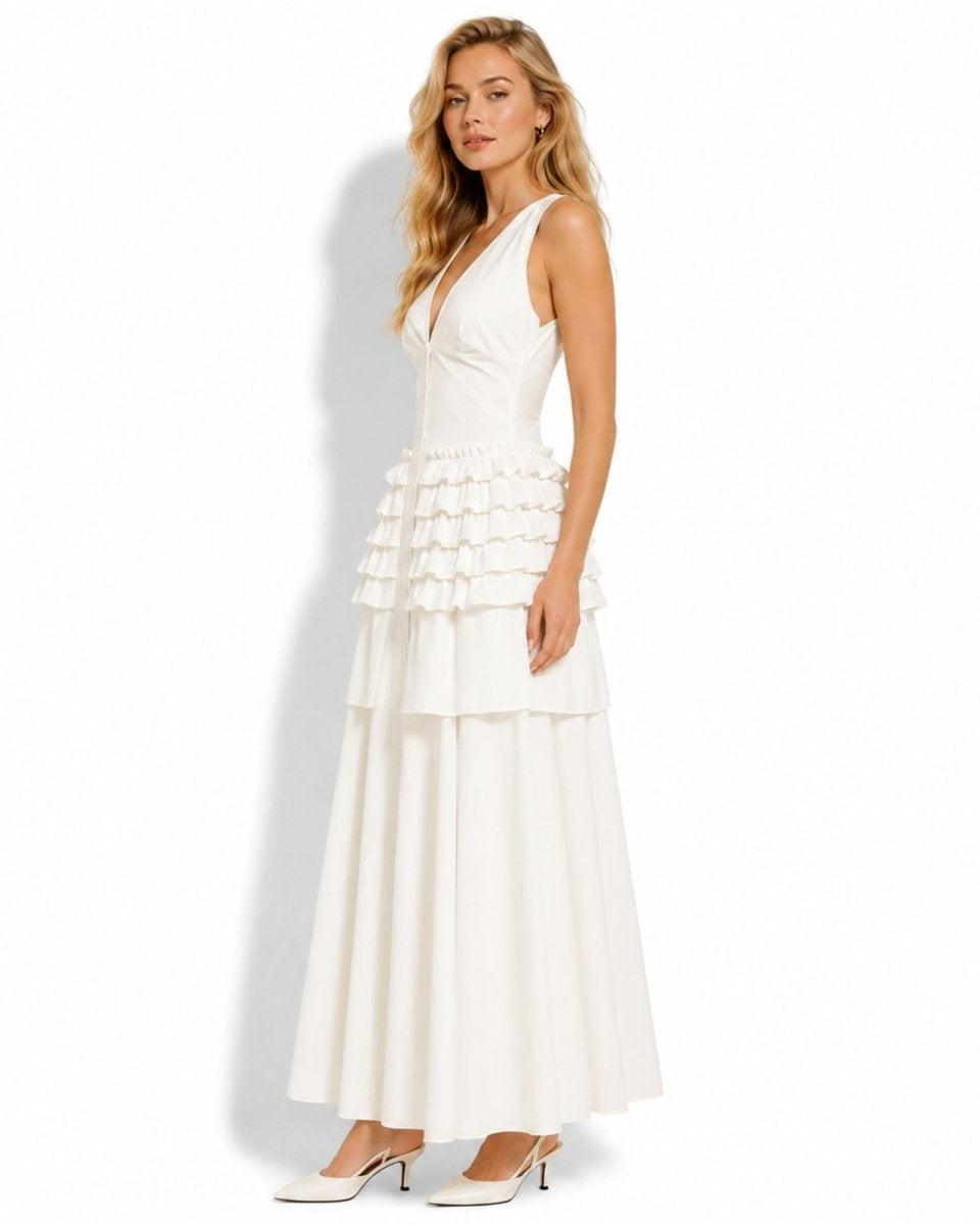 A woman modeling a sleeveless white maxi dress with a structured bodice, ruffled tiered waist, and flowing skirt, paired with white pointed-toe heels against a plain white studio background.