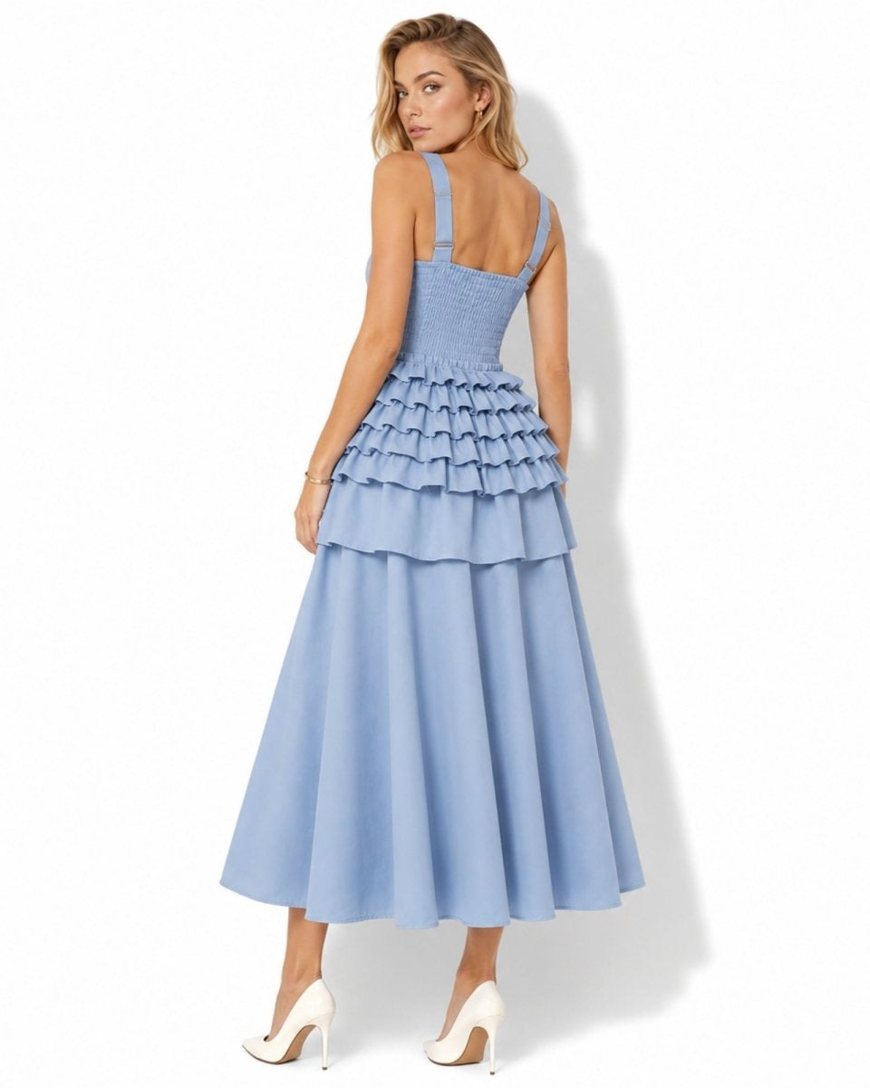 A woman modeling a light blue sleeveless maxi dress with a smocked bodice, tiered ruffle skirt, and white high heels against a white studio background.