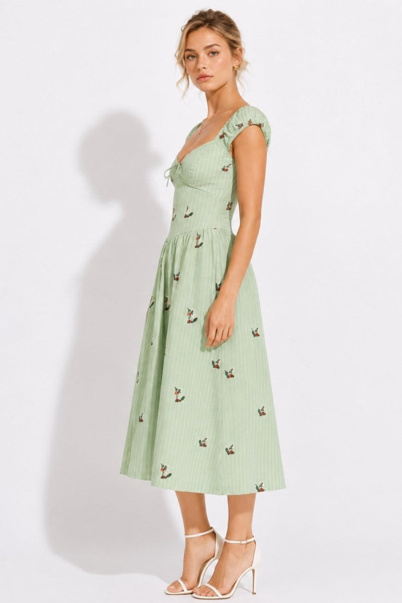 A woman modeling a light sage green midi dress with a small bird print, ruffled cap sleeves, and a smocked bodice, paired with white strappy high heels against a white studio background.