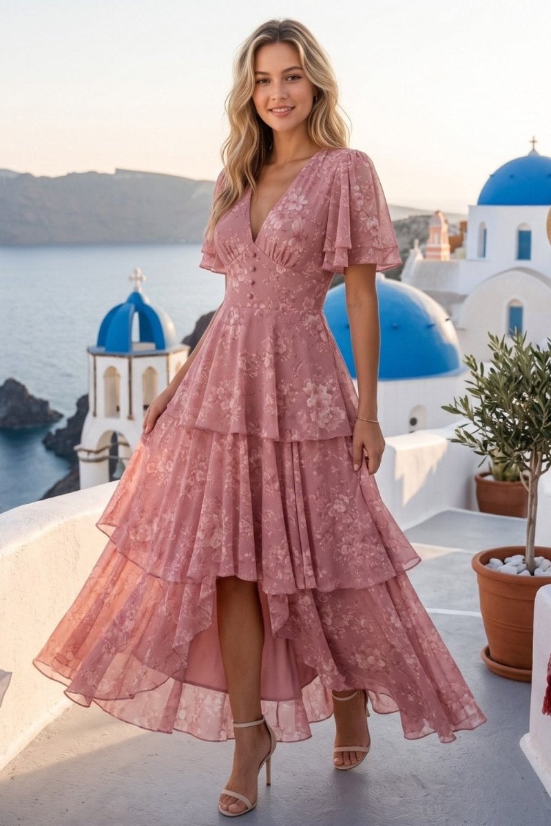A woman stands on a white balcony overlooking the sea with blue-domed buildings in the background, wearing a dusty pink tiered lace dress with flutter sleeves and high-low hem.