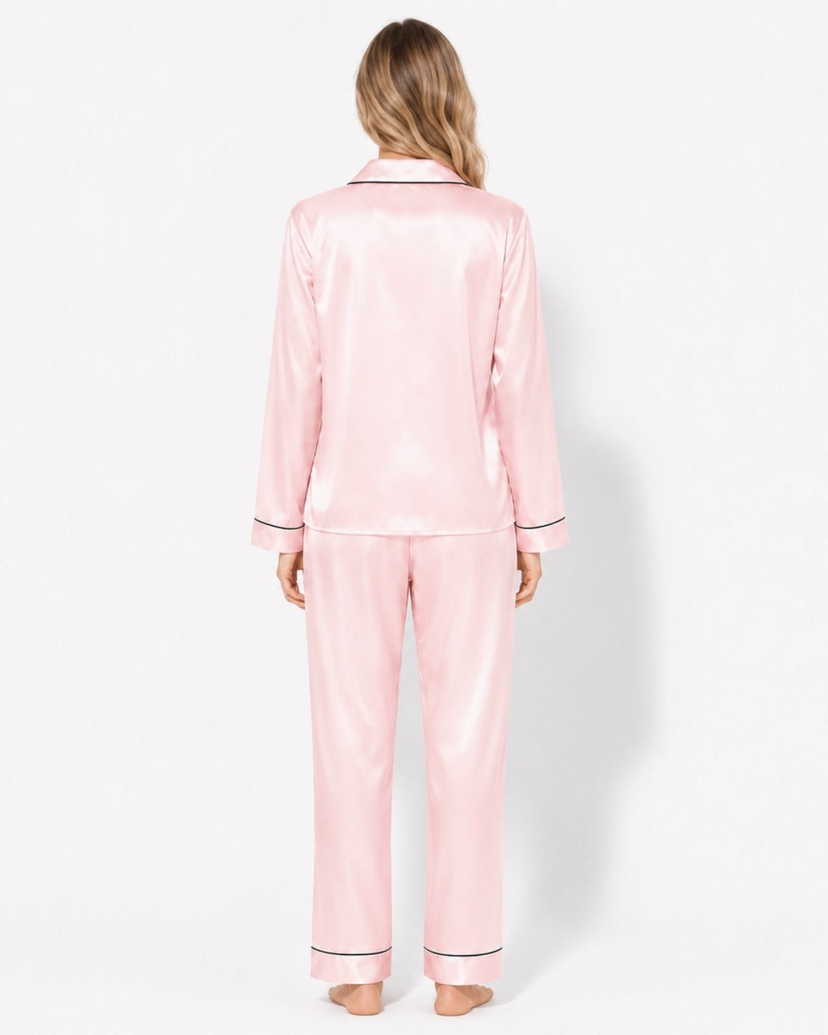 A woman viewed from behind wearing a light pink satin pajama set with thin dark piping trim on the collar, cuffs, and hems, against a plain white studio background.