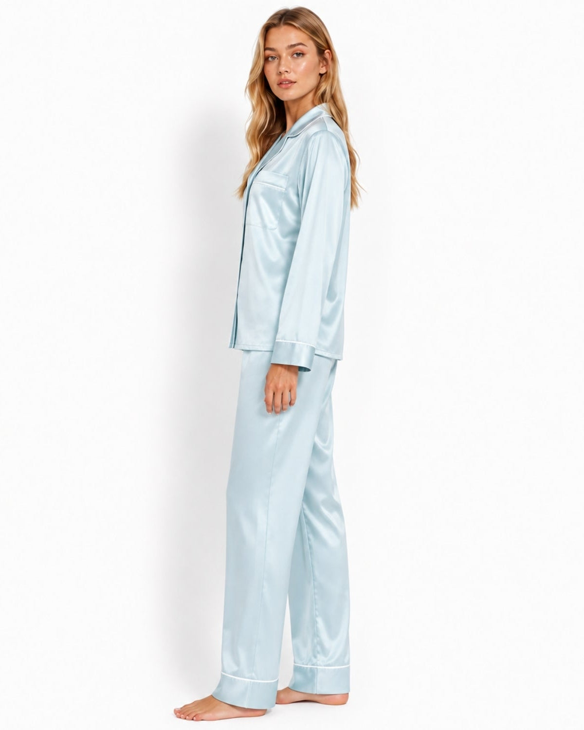 A woman modeling a light blue satin pajama set consisting of a long-sleeve button-up shirt and matching pants, standing against a plain white studio background.