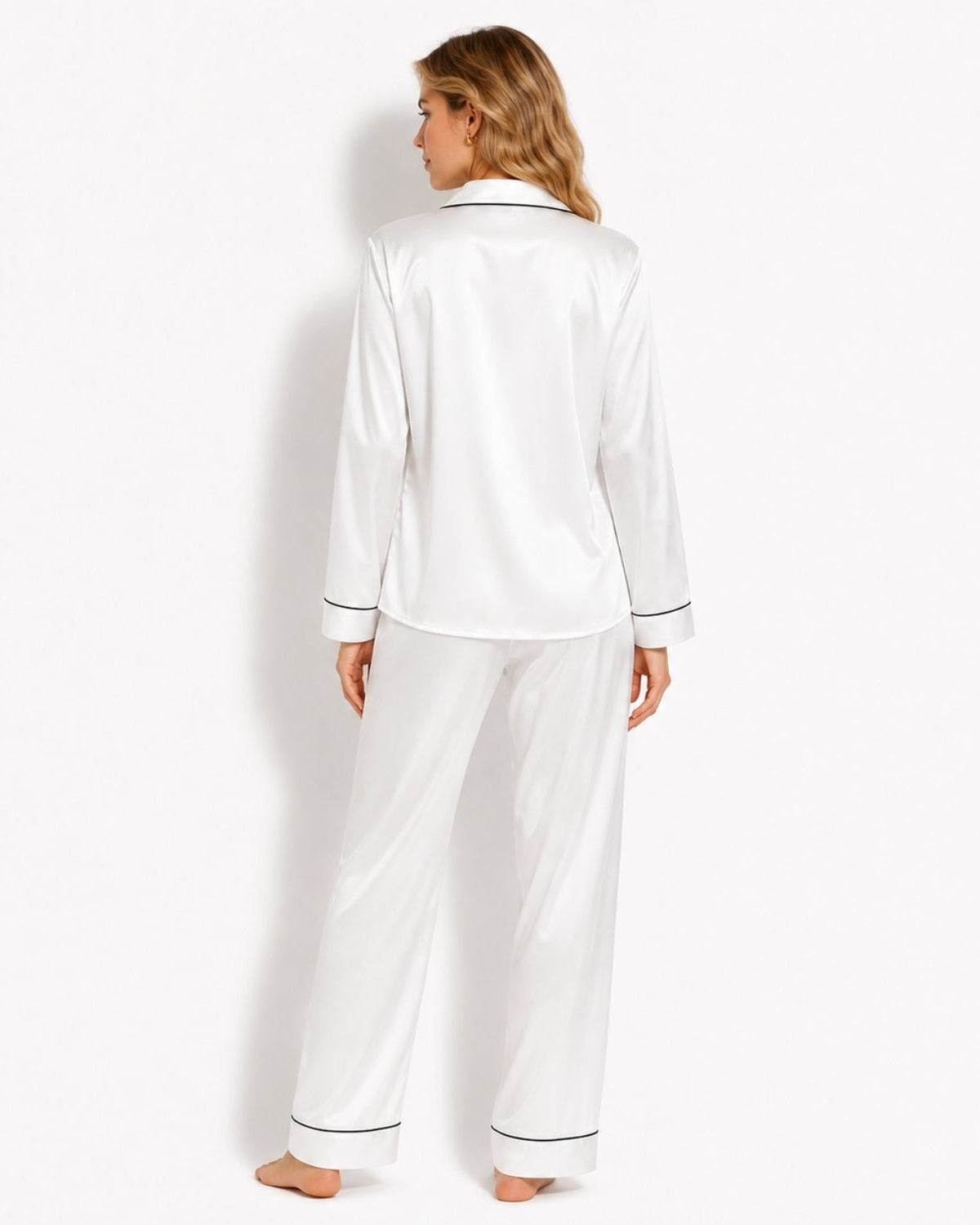 A woman stands barefoot against a plain white studio background, viewed from the back, wearing a white satin pajama set with thin dark piping trim along the collar, cuffs, and pant hems.