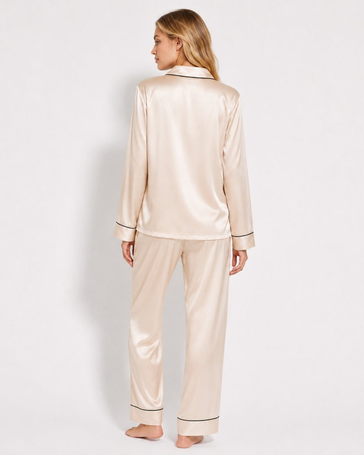 A woman stands barefoot against a plain light background wearing a satin pajama set with a long-sleeve button-front top and matching pants. The pajamas are a warm off-white/cream with thin dark piping trim.