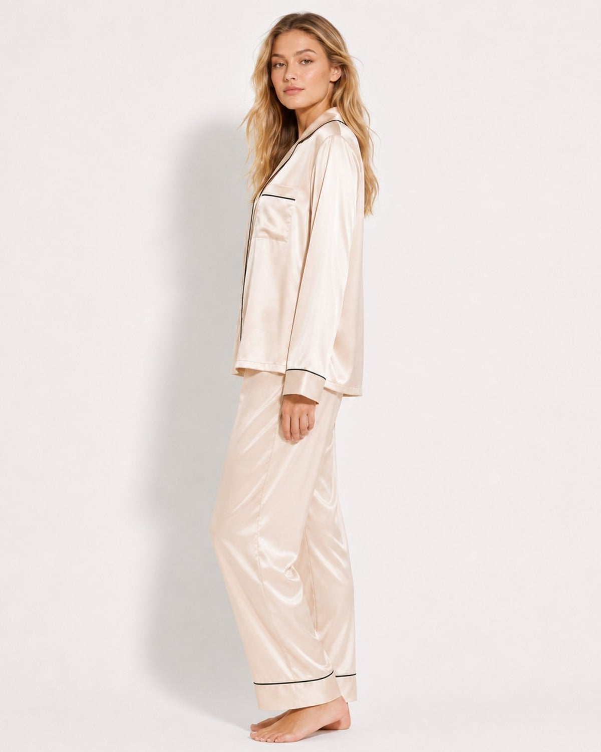 A woman modeling a satin pajama set consisting of a long-sleeve button-up top and matching wide-leg pants, in a light champagne beige color with thin dark piping details, set against a plain white studio background.