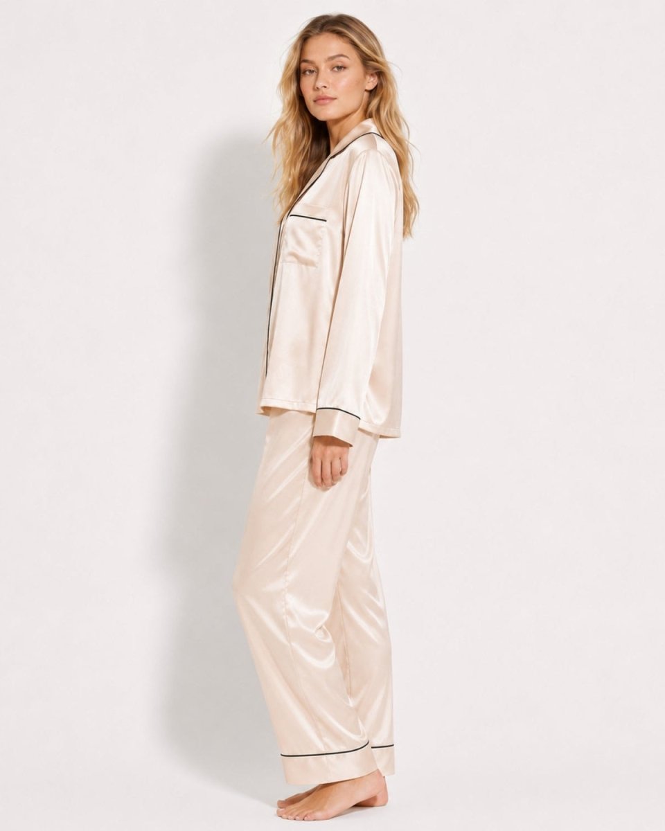 A woman modeling a satin pajama set consisting of a long-sleeve button-up top and matching wide-leg pants, in a light champagne beige color with thin dark piping details, set against a plain white studio background.