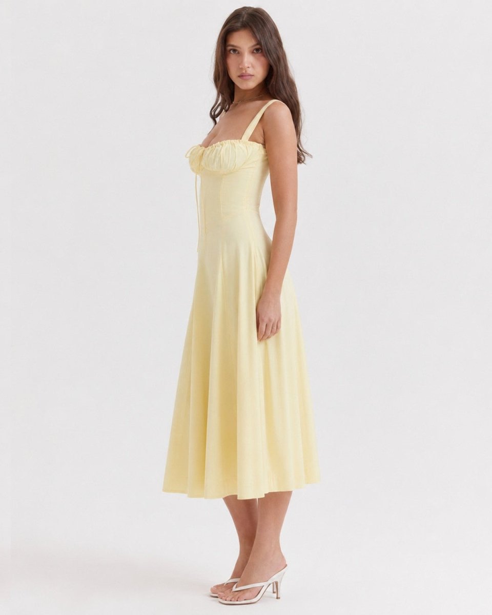 A woman modeling a pale yellow midi dress with thin straps, a gathered sweetheart neckline, and a flared skirt, photographed against a plain light gray studio background.