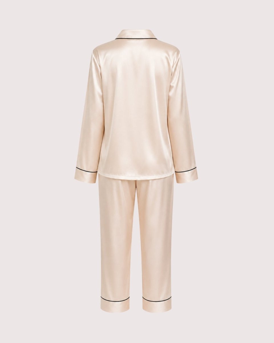 A two-piece satin pajama set in a light champagne beige color, shown from the back. The set includes a long-sleeve top with thin black piping along the collar, cuffs, and hem, and matching straight-leg pants with black piping at the cuffs.
