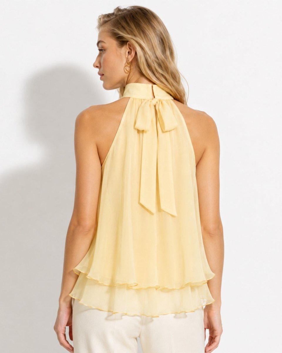 A woman shown from the back wearing a pale yellow halter top with a large bow at the neck and a layered ruffle hem, paired with light pants against a plain white studio background.
