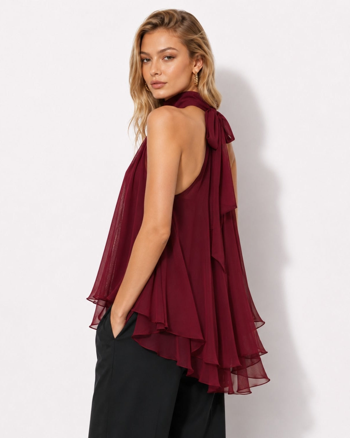 A woman modeling a sleeveless burgundy ruffled top with a tie-neck detail and black pants, photographed against a plain white studio background.