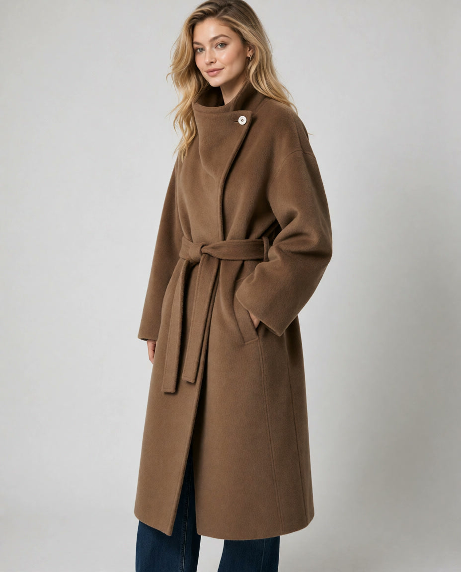 A woman modeling a mid-length  amber belted coat with an asymmetrical button detail at the collar, paired with dark blue jeans, against a neutral light gray studio background.