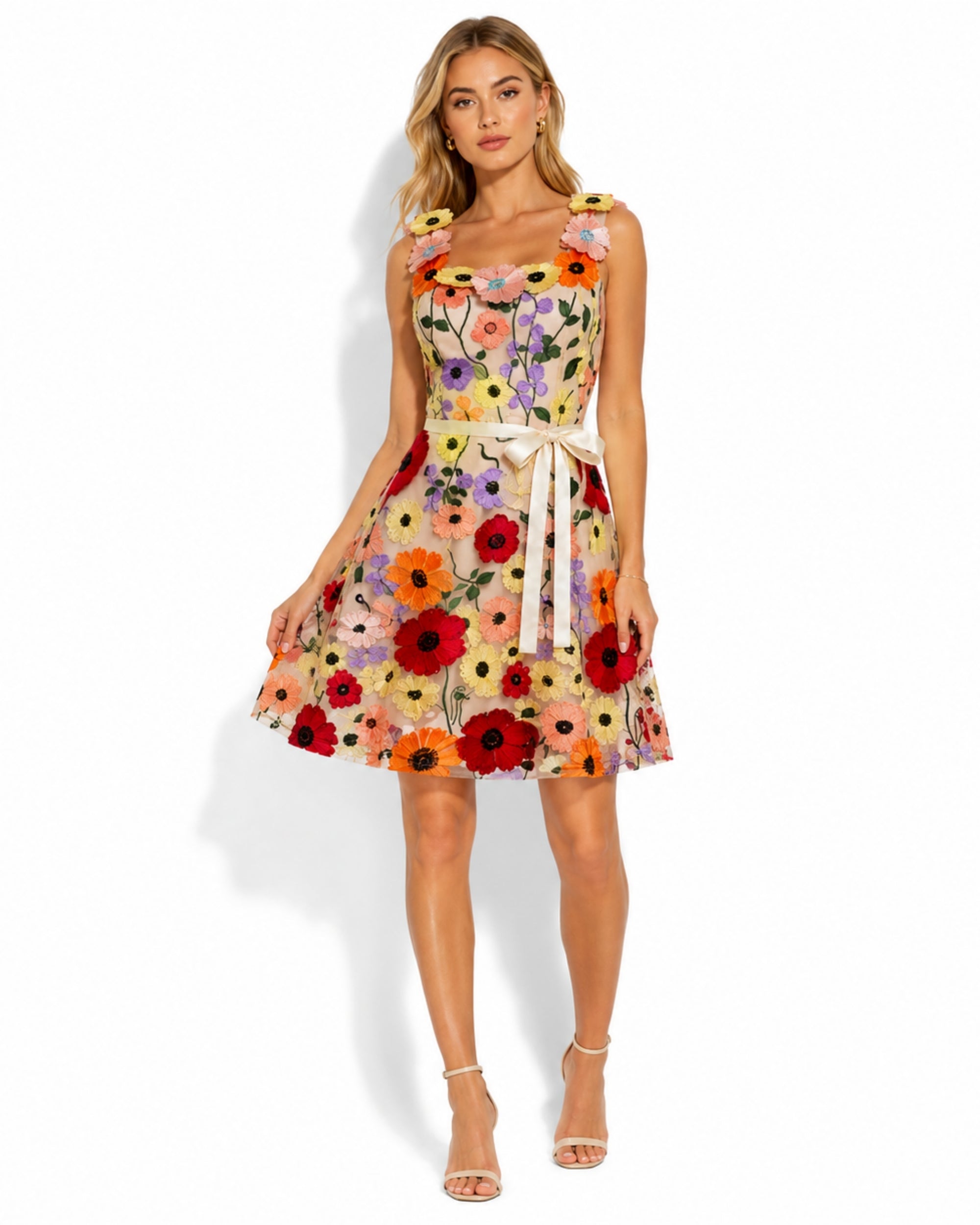 A woman modeling a sleeveless fit-and-flare dress with a colorful floral print, ruffled shoulder straps, a tie waist, and a flared skirt, photographed against a clean white studio background.