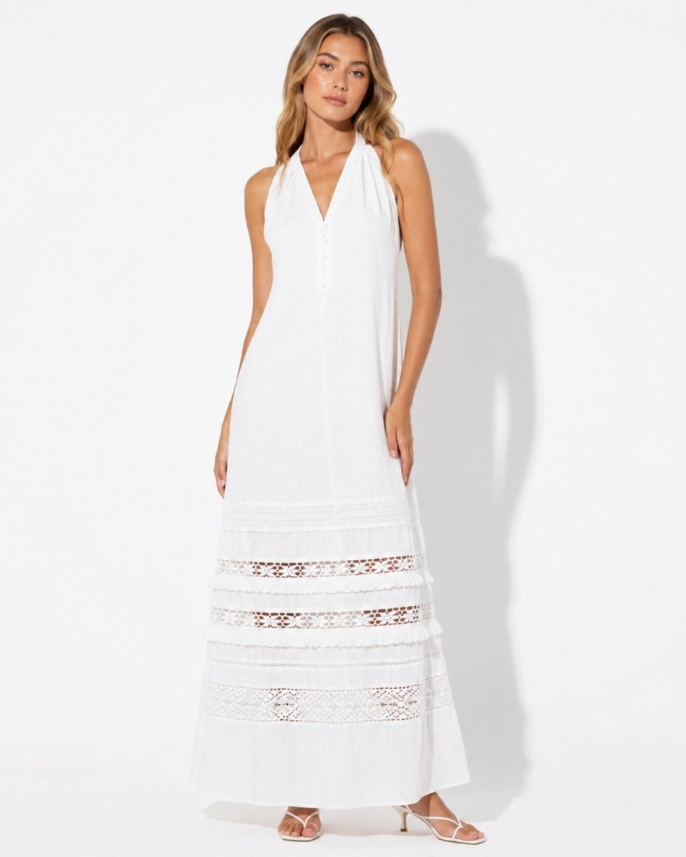 Woman modeling a flowy white halter maxi dress with lace tiered cutouts on a seamless white background.