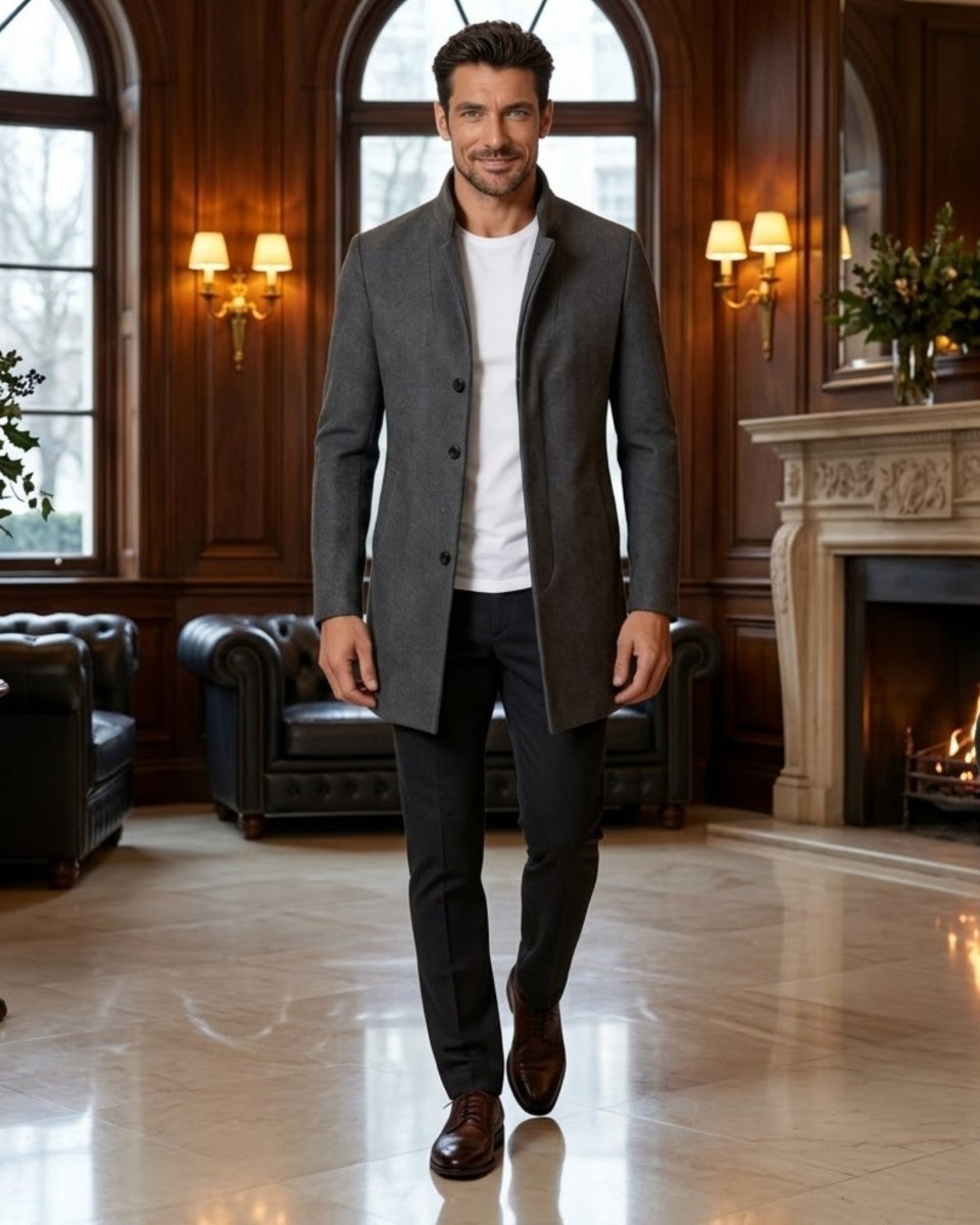A man walking in a grand, elegant room with wood-paneled walls, a fireplace, and leather seating, wearing a dark gray mid-length coat over a white t-shirt, dark trousers, and brown dress shoes.
