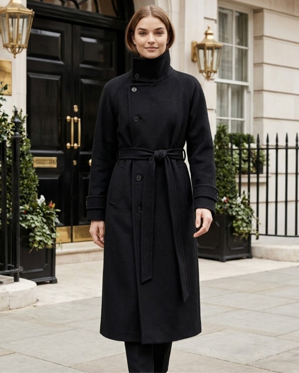 A woman wearing a long black wrap coat with a high mock neck and waist tie, standing in front of a classic London-style black door and stone building facade.
