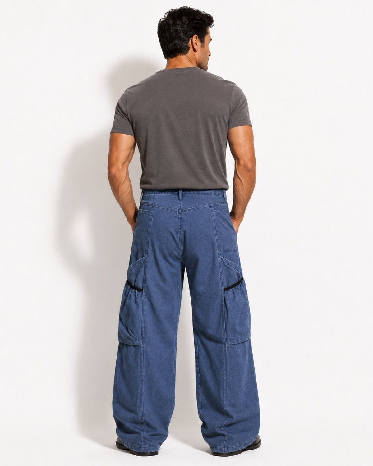A man viewed from behind wearing wide-leg blue cargo pants and a dark gray t-shirt, standing against a plain white studio background.