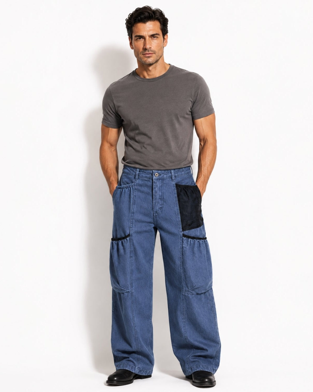 A man modeling a pair of wide-leg blue denim pants with black contrast patch pockets, paired with a dark gray t-shirt and black shoes, standing against a plain white studio background.