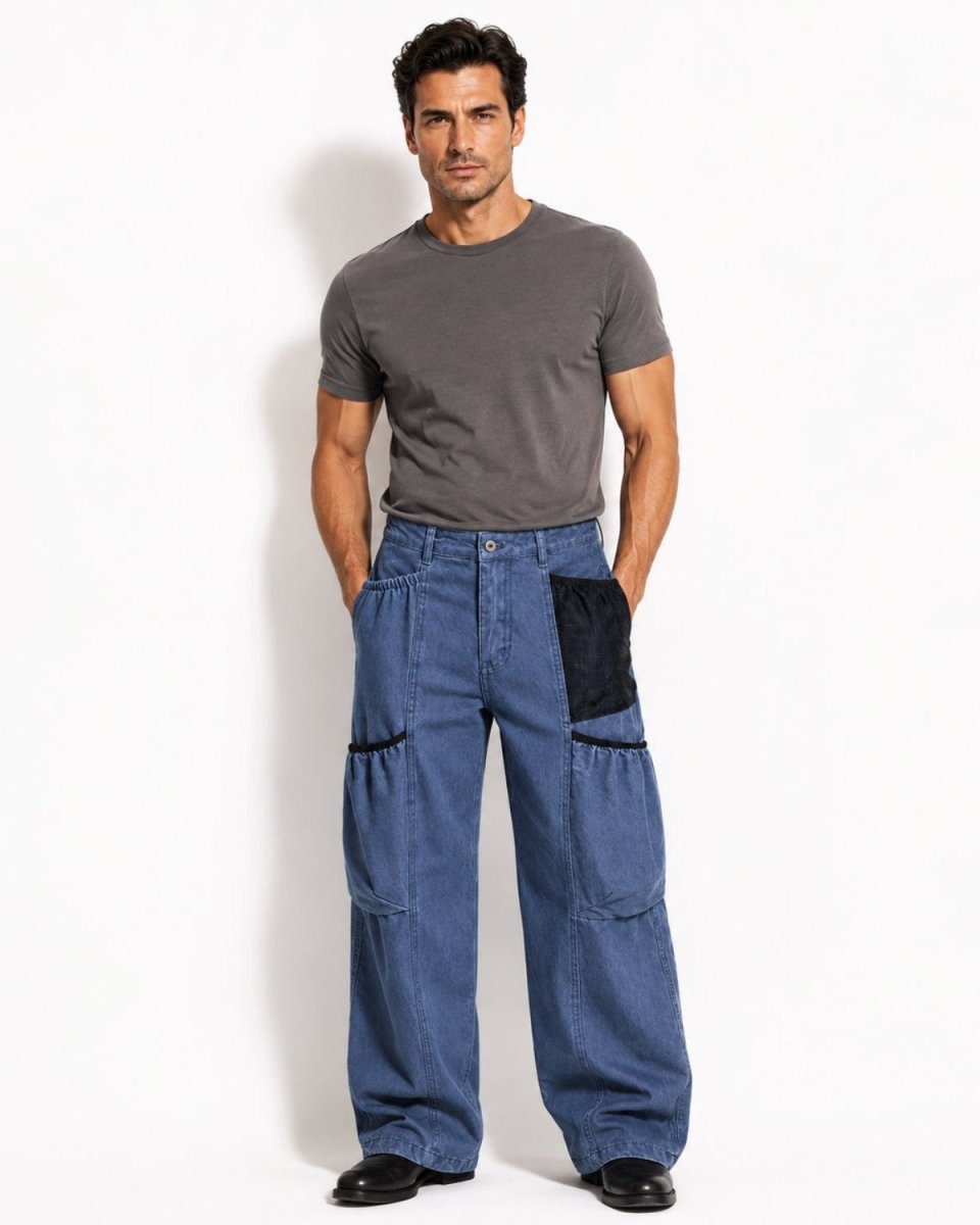 A man modeling a pair of wide-leg blue denim pants with black contrast patch pockets, paired with a dark gray t-shirt and black shoes, standing against a plain white studio background.