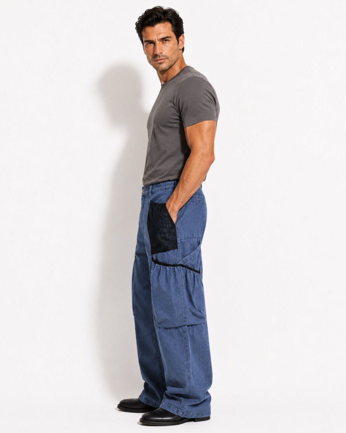 A man wearing blue wide-leg cargo pants with black contrast panels and a dark gray t-shirt, standing in profile against a white studio background.