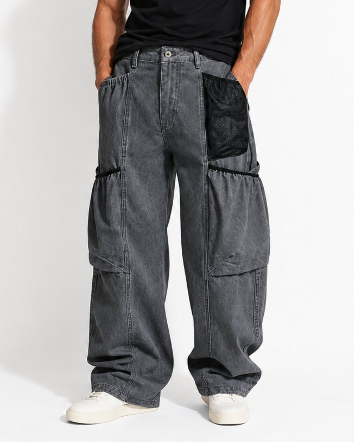 A person wearing very baggy dark gray washed cargo pants with black contrast pocket panels, paired with a black shirt and white sneakers, photographed against a plain light gray studio background.