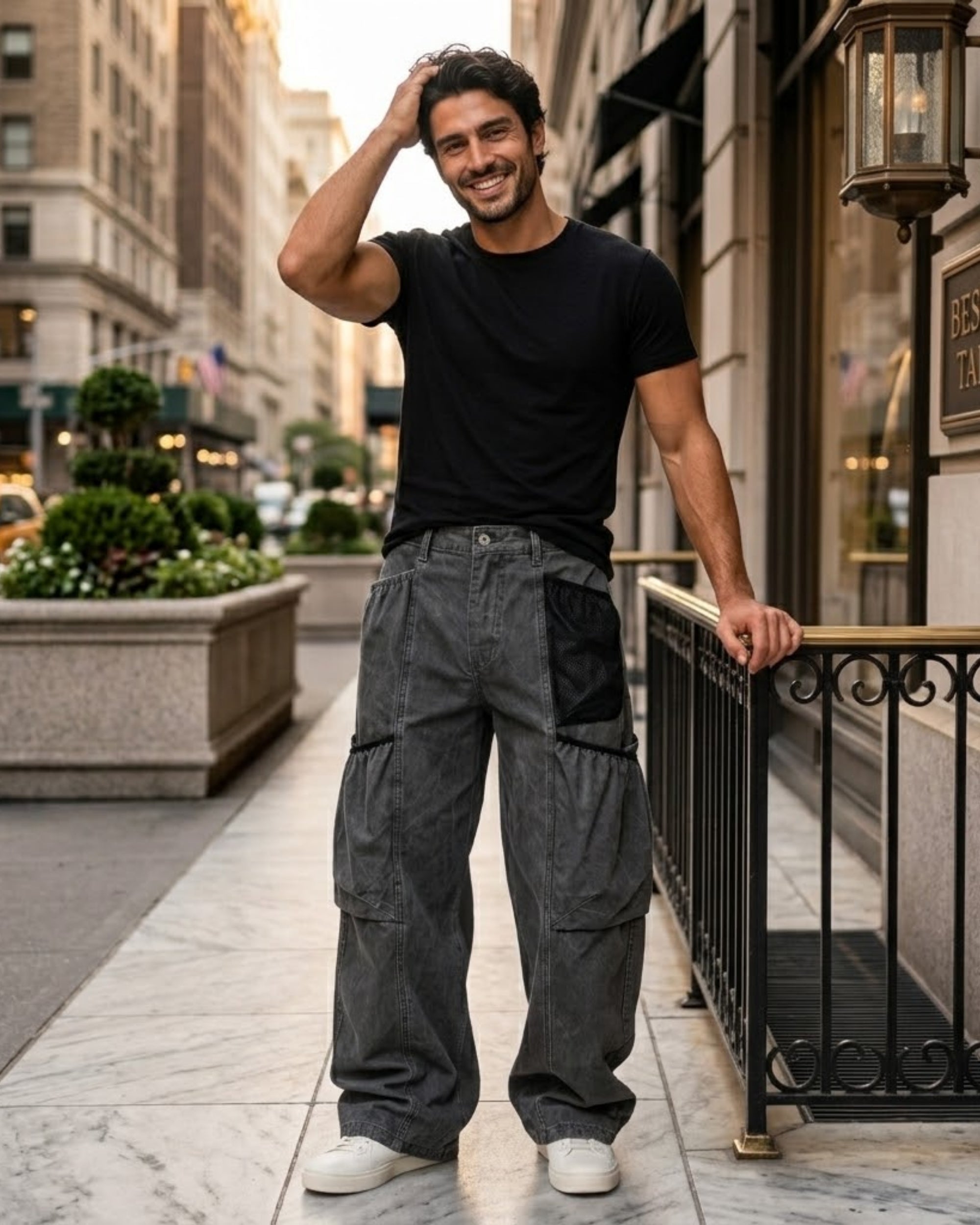A man wearing a black crewneck t-shirt and dark gray wide-leg cargo pants with black side pockets, standing on a city sidewalk next to a building.