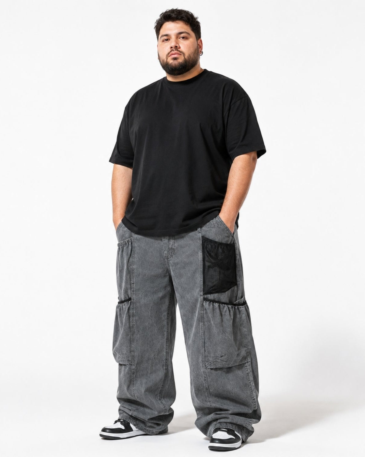 A man modeling a black crewneck t-shirt and wide-leg gray cargo pants with black mesh pocket details, standing against a plain white studio background.