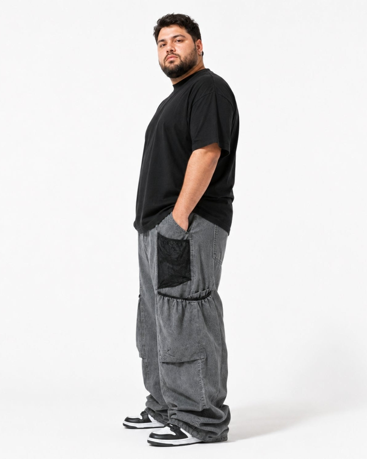 A man wearing a black t-shirt and loose gray cargo pants with black side pockets, standing in profile against a plain white studio background.