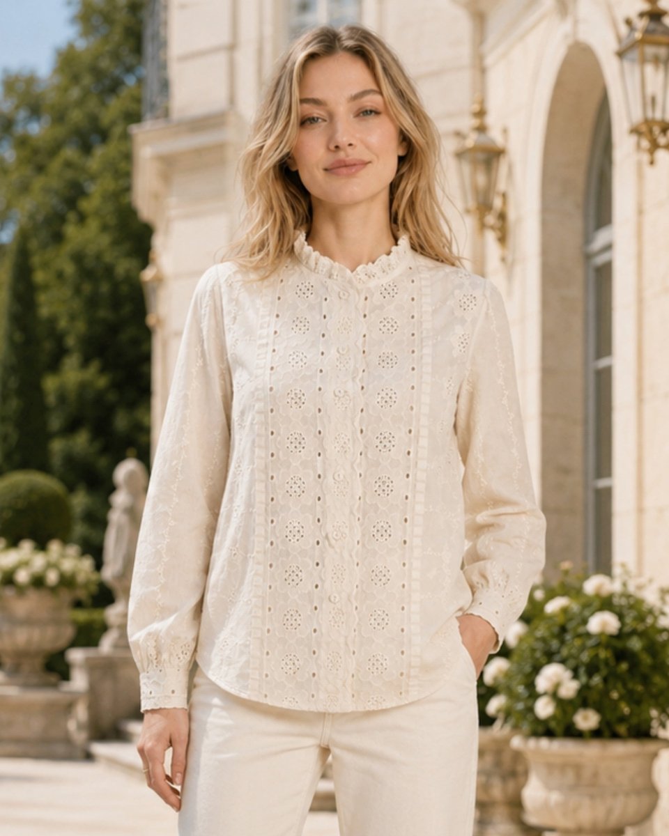 A woman wearing a cream-colored eyelet lace blouse and matching cream trousers, standing outdoors in front of a grand classical building with greenery.