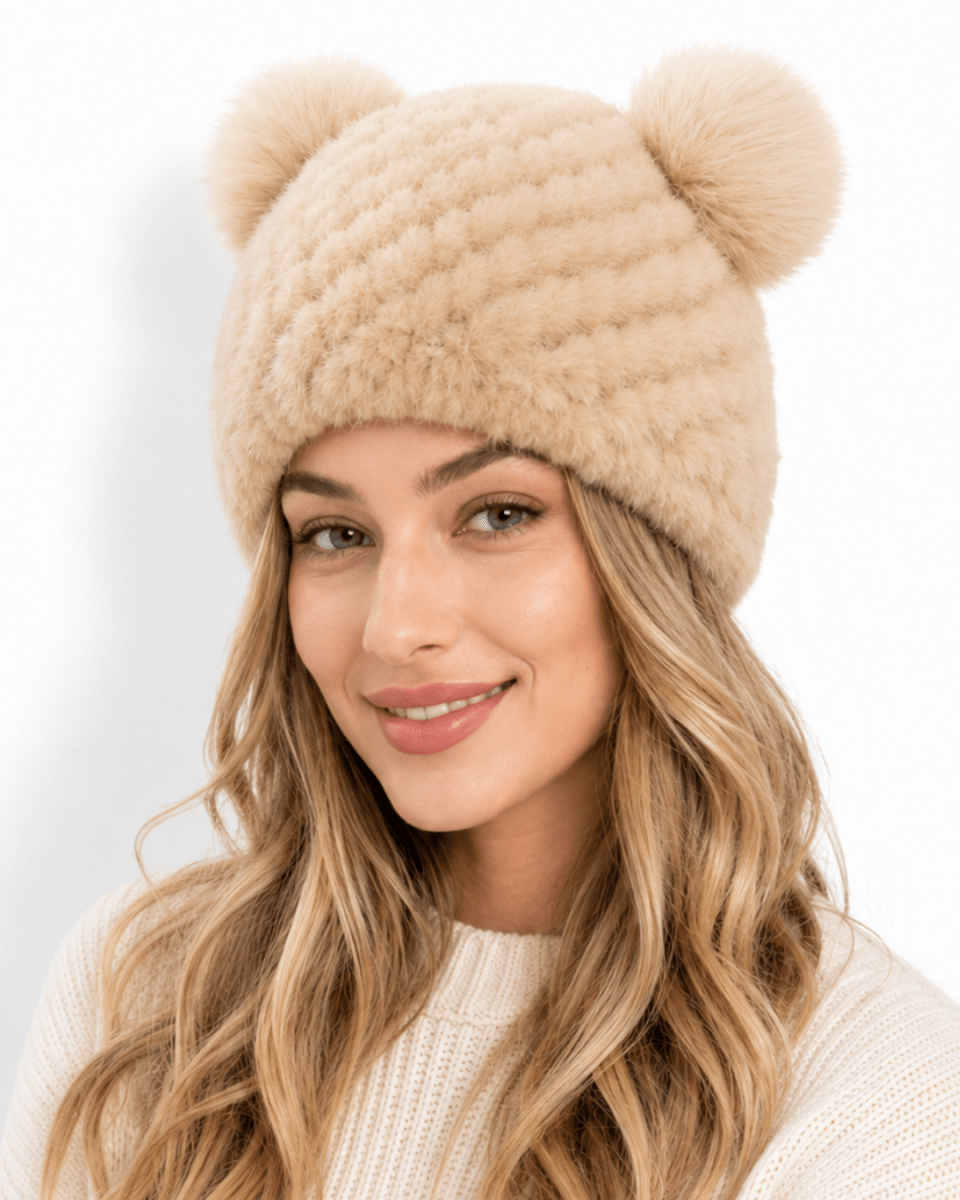 A woman wearing a beige chunky knit beanie with two pom-poms on top, photographed against a clean white background.