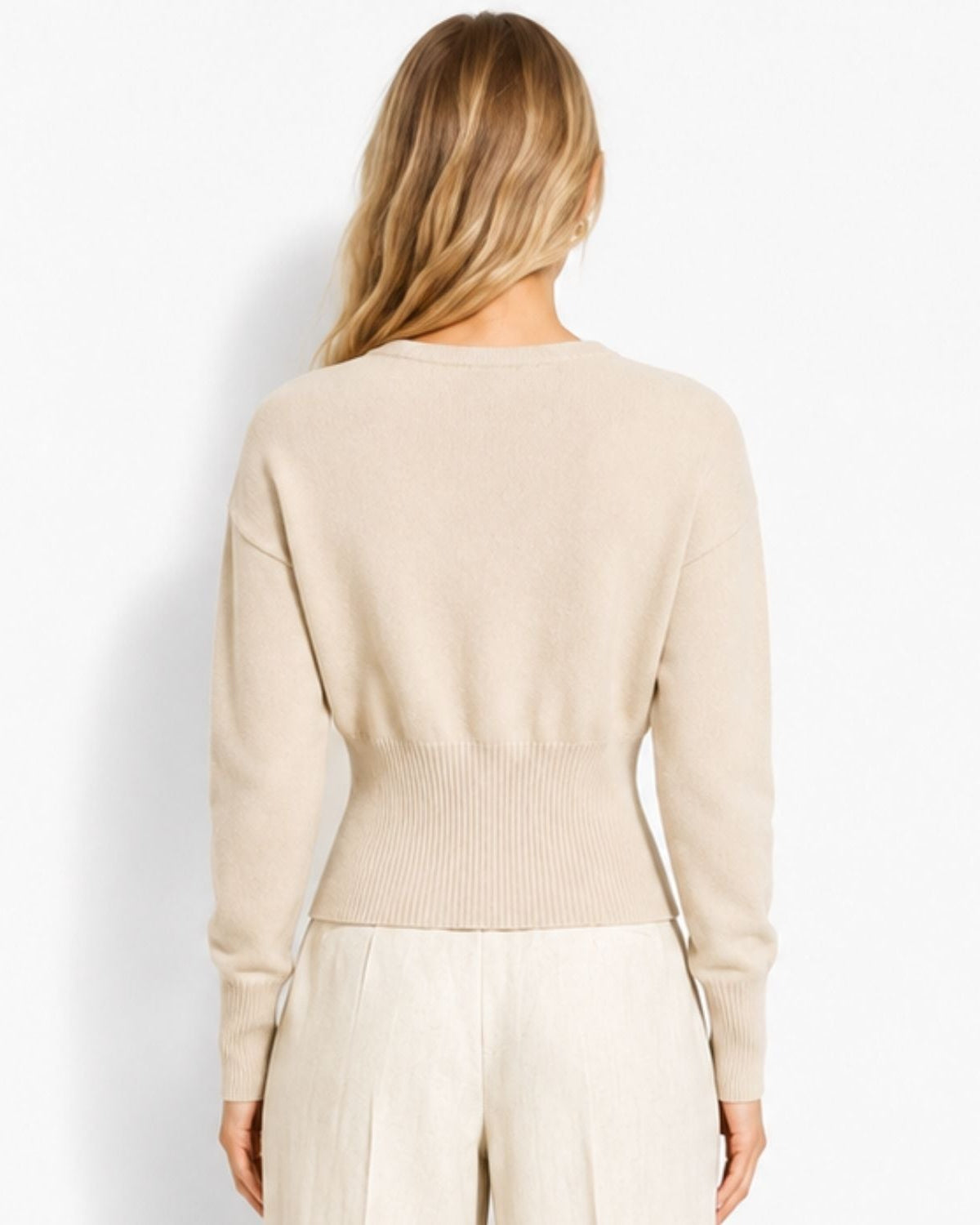 A woman viewed from the back wearing a light beige knit sweater and matching light pants against a plain white studio background.