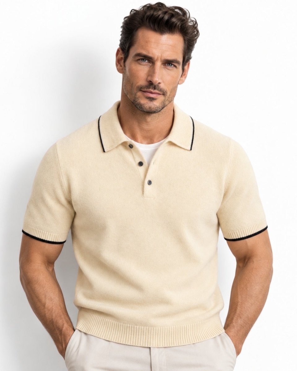 A man wearing a beige-colored short-sleeve knit polo shirt with black trim on the collar and sleeve cuffs, paired with light beige trousers, against a plain white studio background.