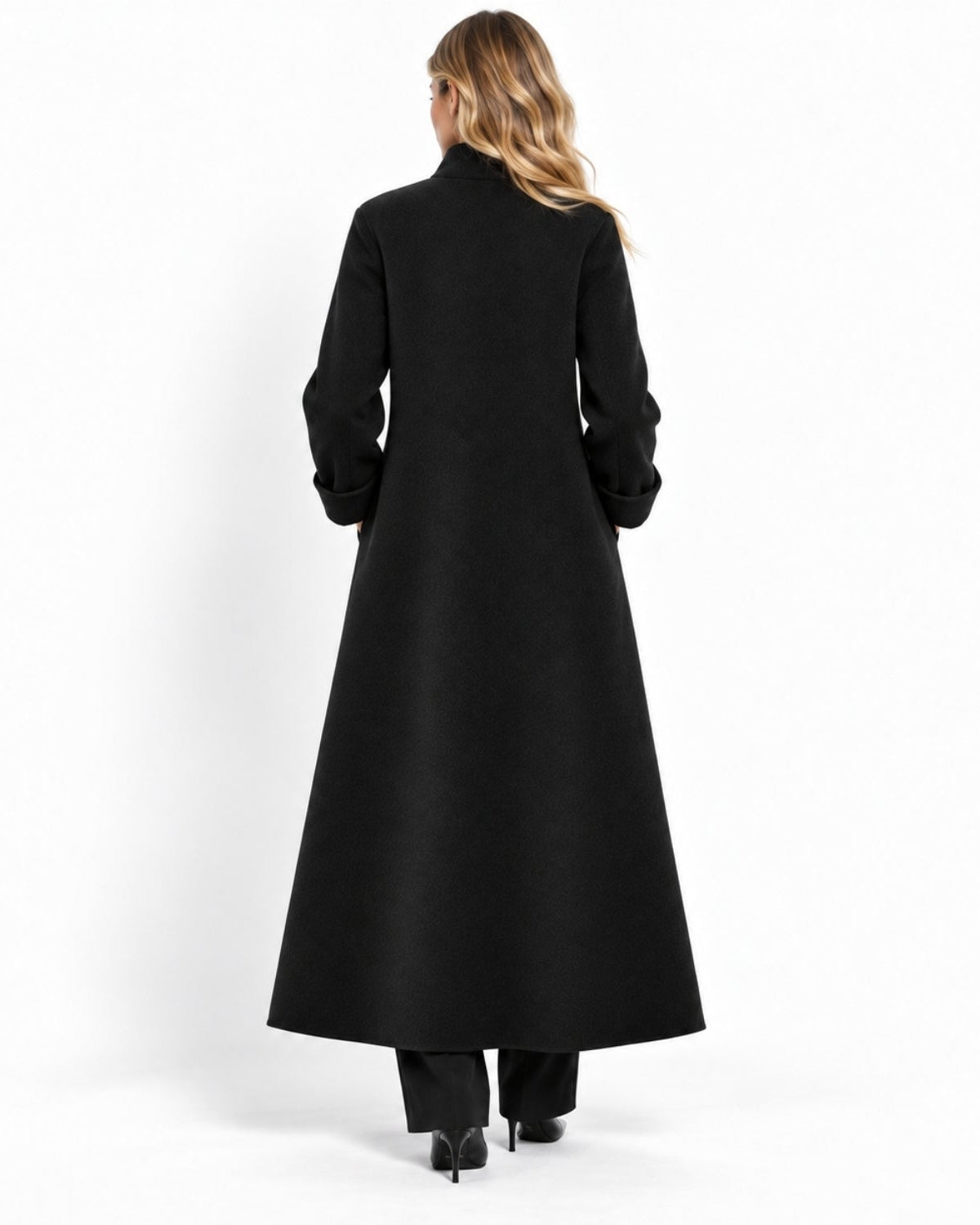 A woman viewed from behind wearing a long black coat, black trousers, and black high heels against a plain white studio background.