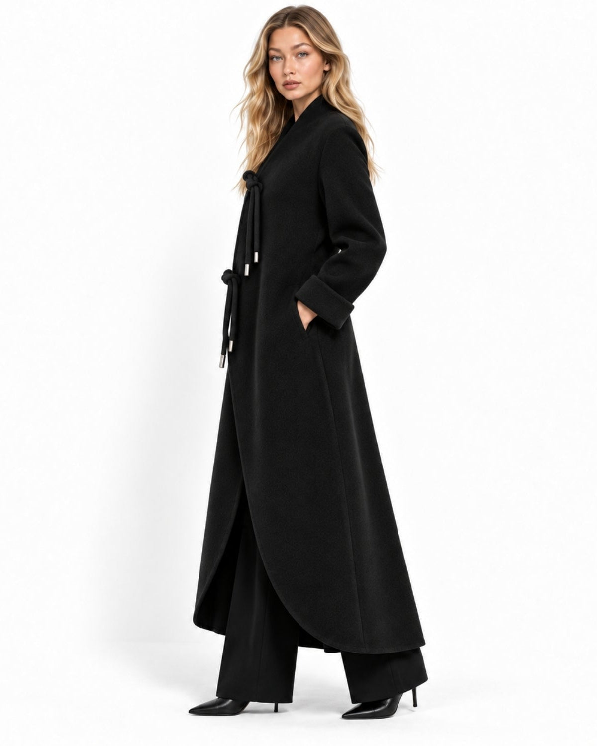 A woman modeling a long black wrap-style coat with a tie belt, wide-leg trousers, and black pointed-toe heels against a plain white studio background.