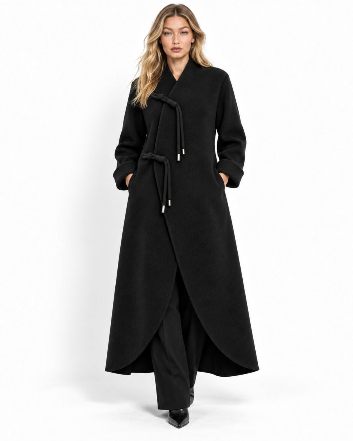 A woman modeling a long black wrap-style coat with a tie belt, black wide-leg trousers, and black shoes against a plain white studio background.