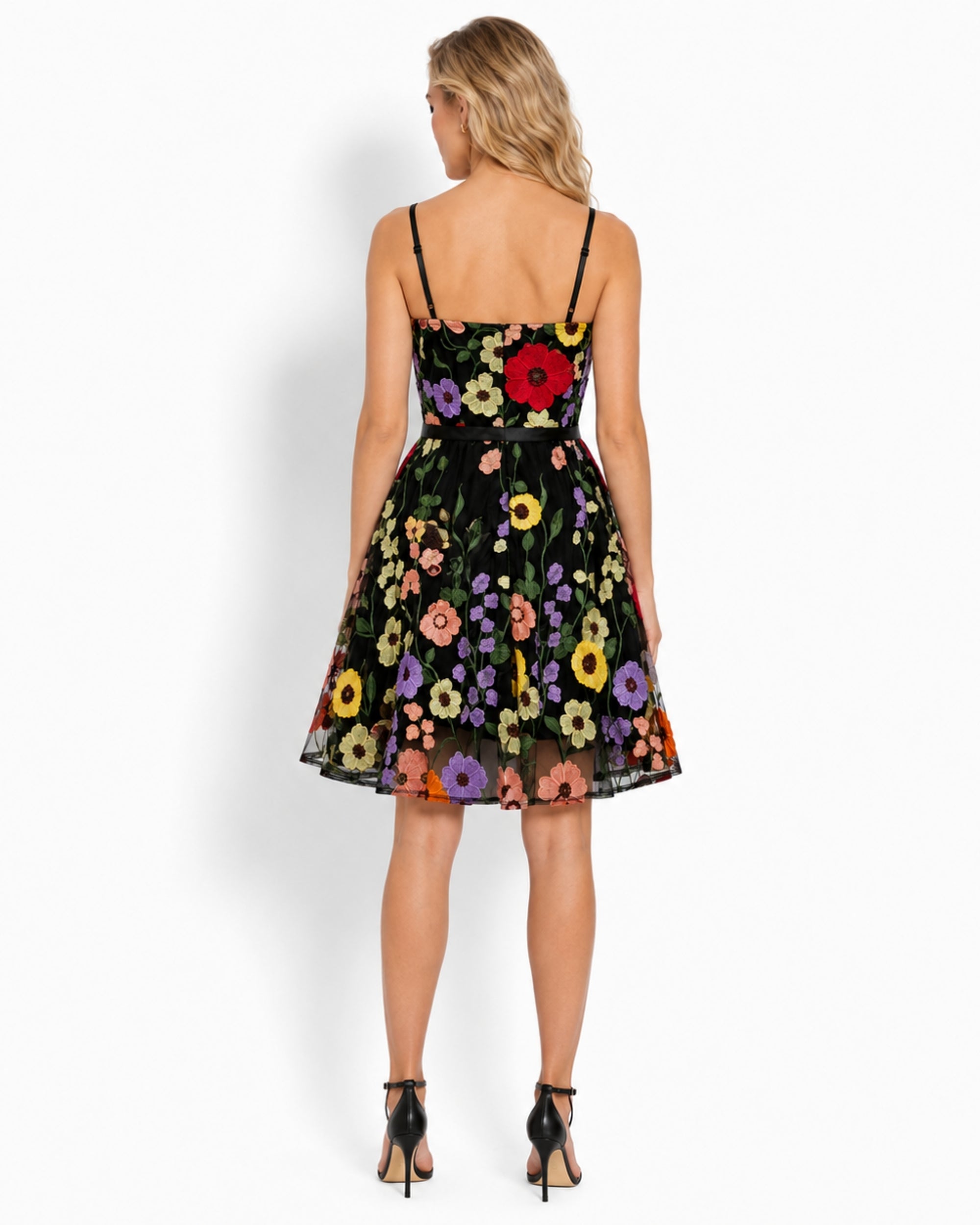 A woman shown from the back wearing a black spaghetti-strap mini dress with a vibrant multicolor floral print, paired with black high heels, against a plain white studio background.
