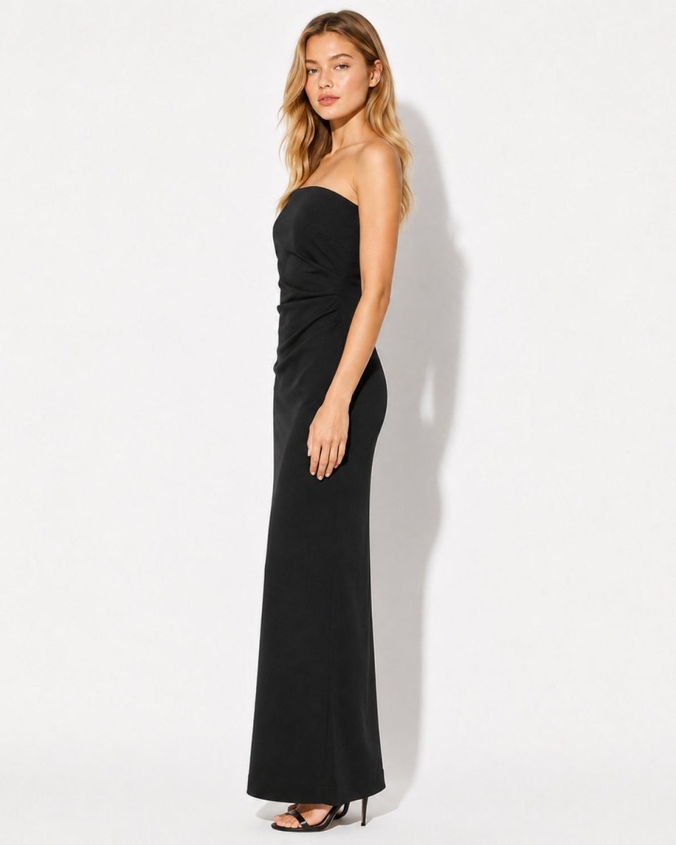 A woman modeling a strapless black maxi dress with a ruched bodice and full-length skirt, standing against a plain white studio background.