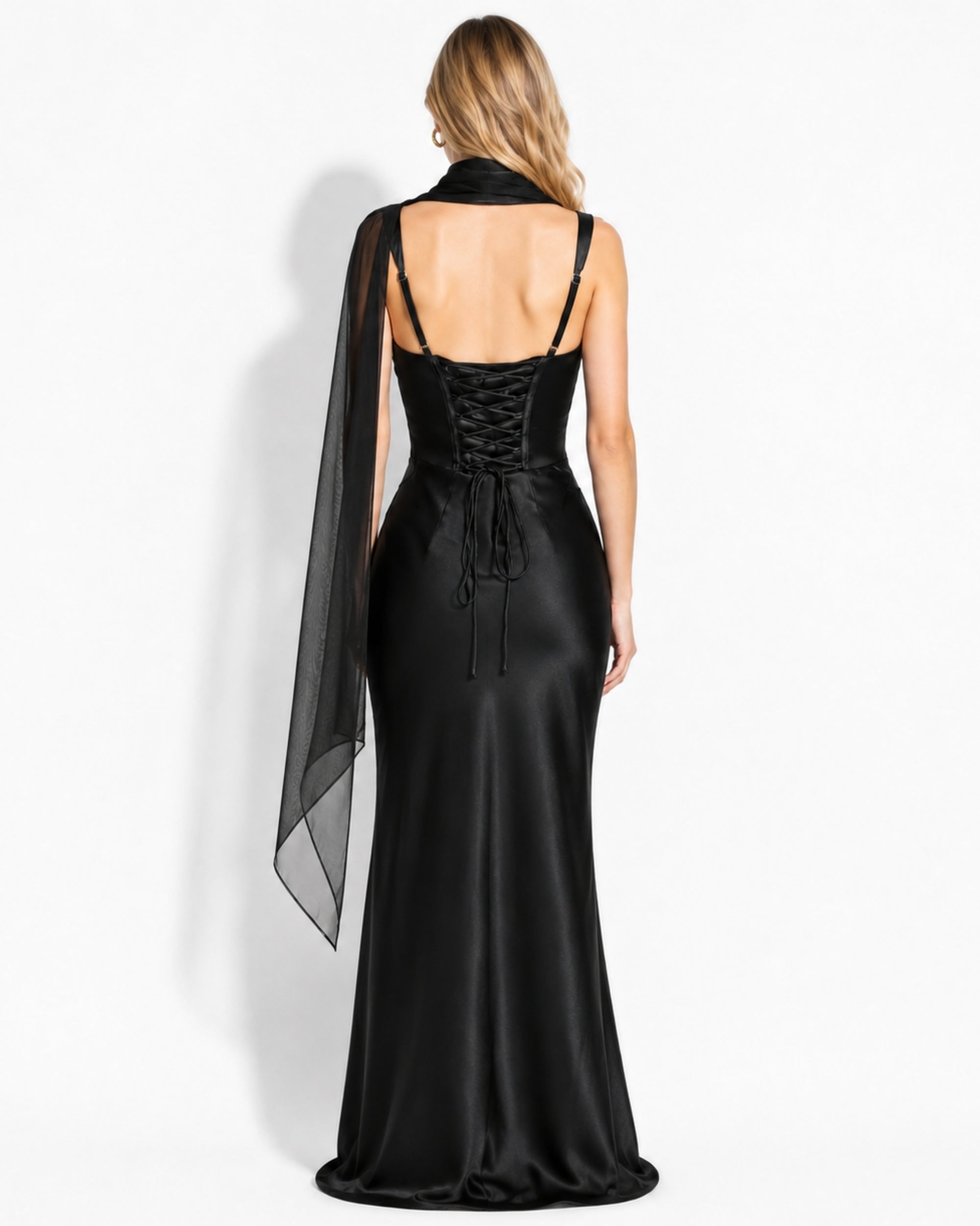 A woman shown from the back wearing a floor-length black satin gown with a lace-up open back and a sheer black draped cape detail, photographed against a plain light background.