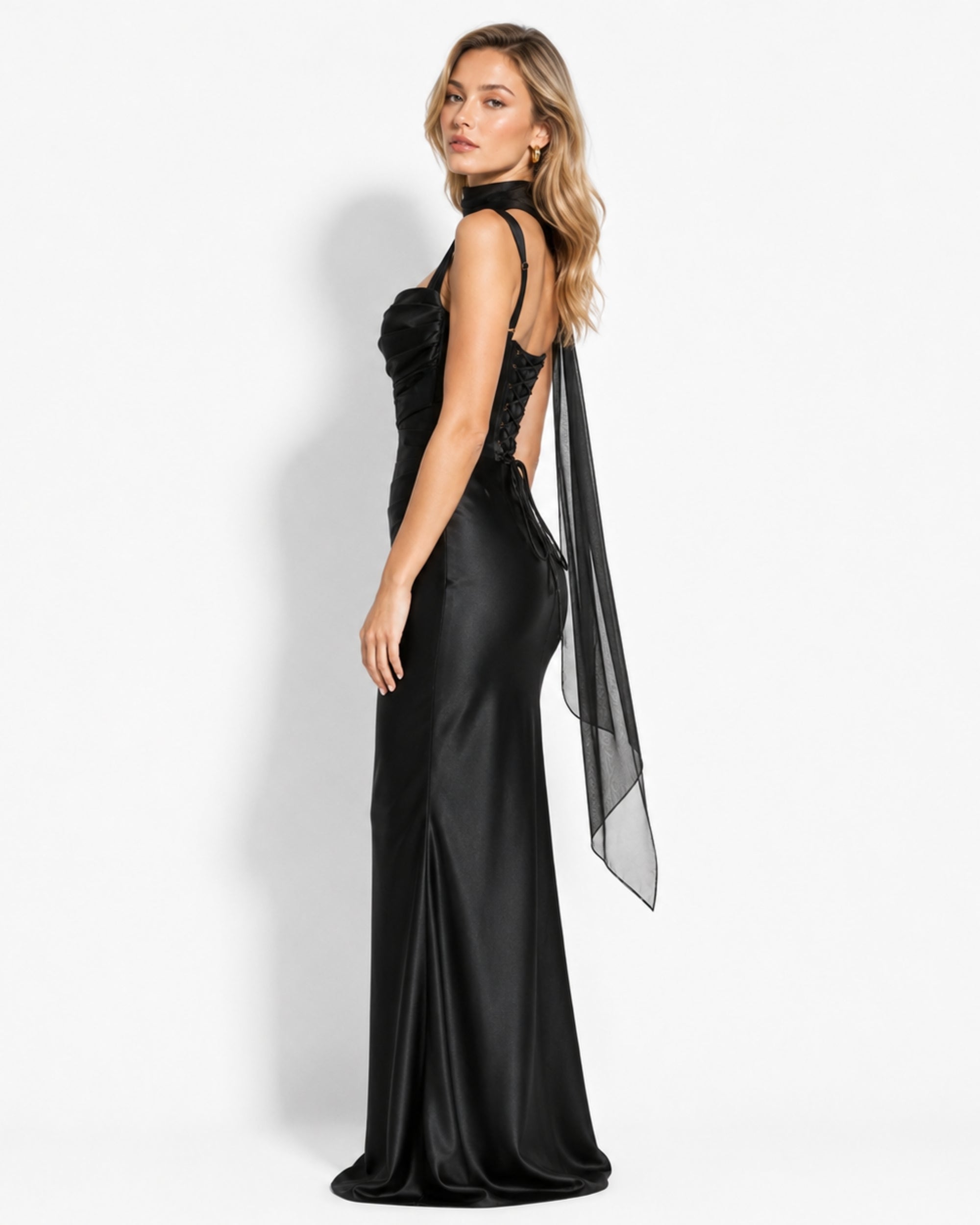 A woman modeling a floor-length black evening gown with a high neckline, lace-up open back, and a sheer draped overlay, photographed against a clean white studio background.