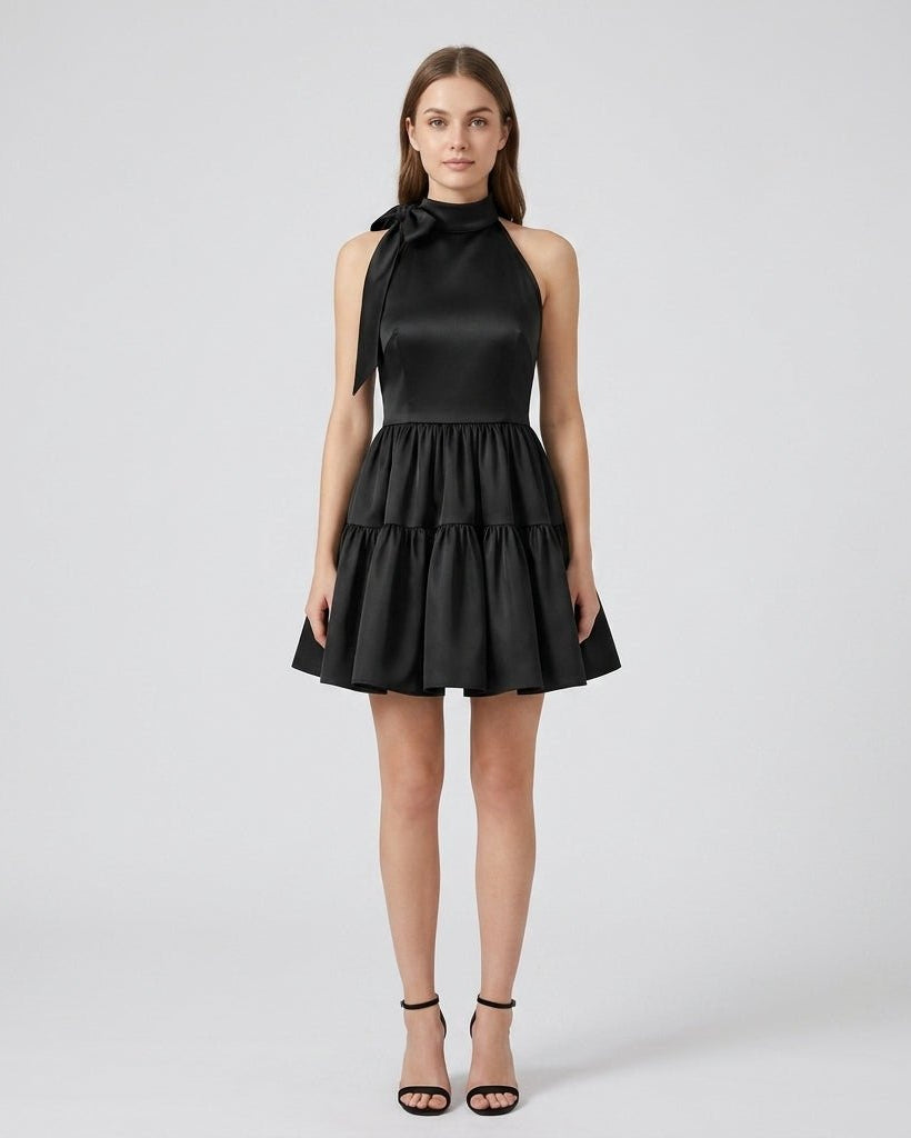 Front view of blonde model wearing the black halter tiered mini dress with bow tie neck and voluminous A-line skirt for cocktail parties.