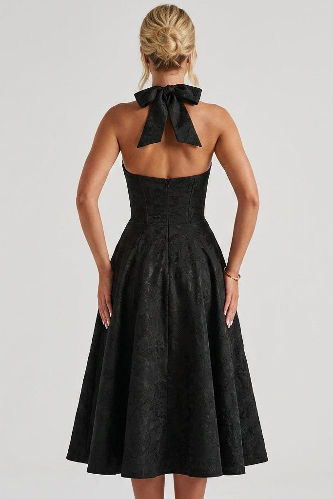 Back view of black fit and flare midi dress showing oversized halter bow tie and open back design.