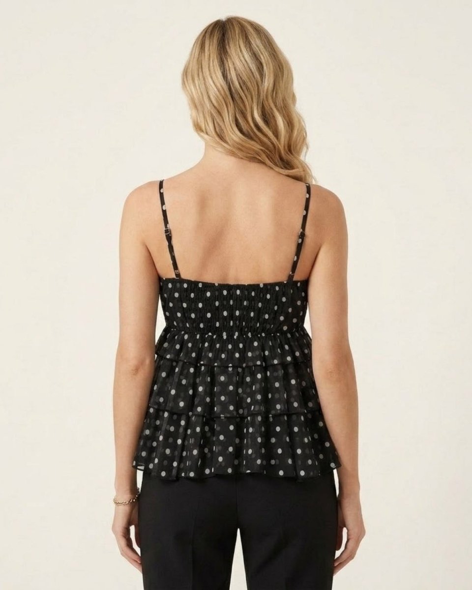 A woman viewed from the back wearing a black ruffled spaghetti-strap top with a small white polka dot pattern, paired with black pants, against a plain off-white background.