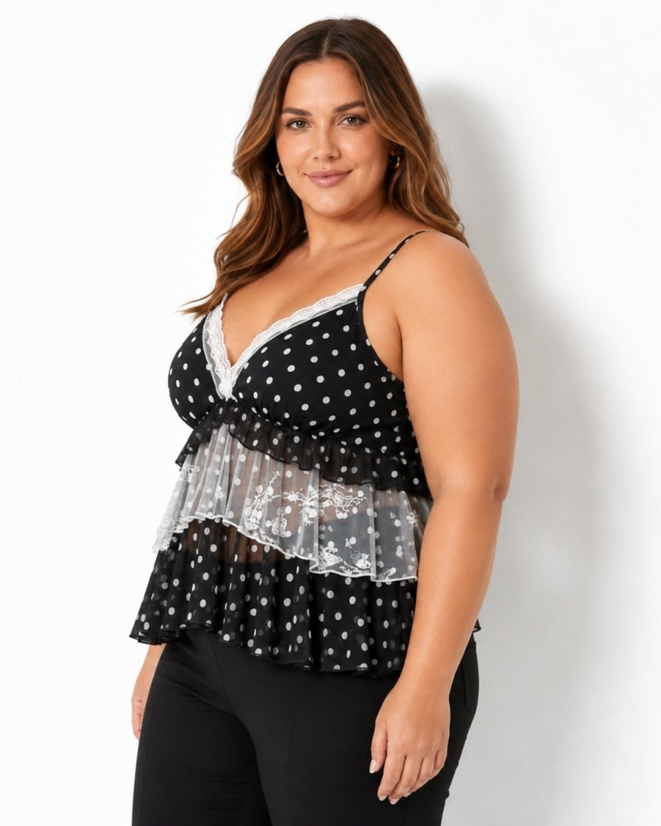 A woman modeling a black camisole top with white polka dots, sheer white lace ruffle layers, and white trim on a solid white background.