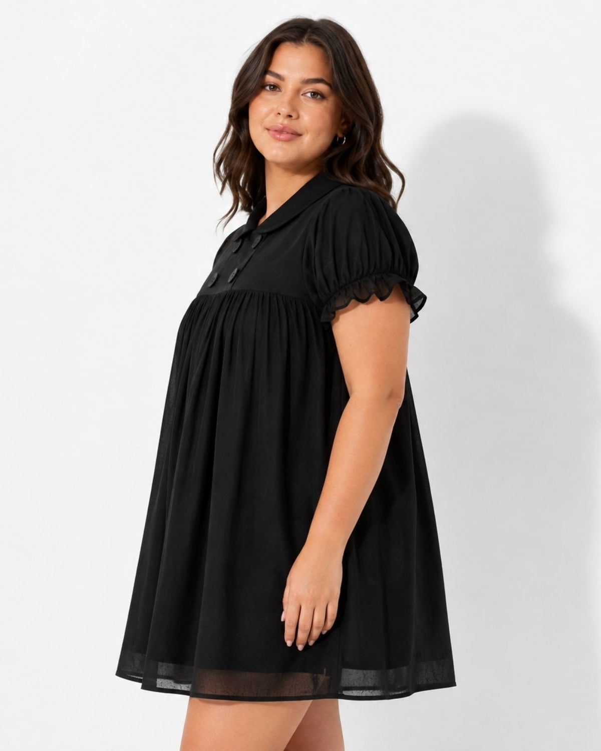 A woman modeling a black short-sleeve babydoll dress with puff sleeves, a gathered waist, and a sheer mesh hem against a white studio background.