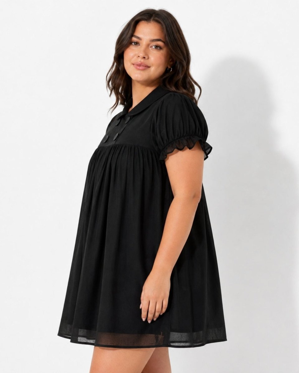 A woman modeling a black short-sleeve babydoll dress with puff sleeves, a gathered waist, and a sheer mesh hem against a white studio background.