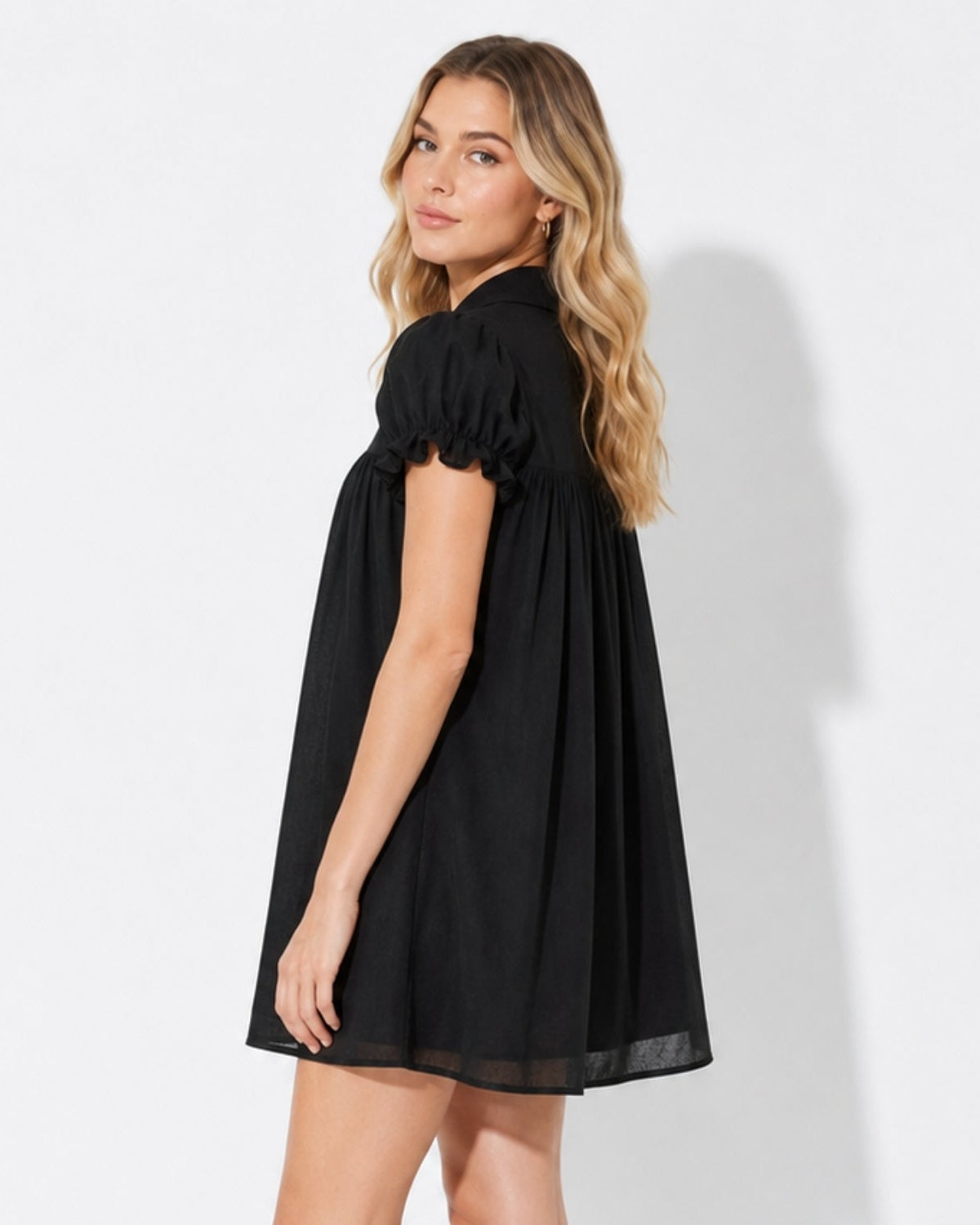 Side profile of a model wearing a voluminous black smock mini dress featuring ruffled short puff sleeves and a flowing A-line silhouette.