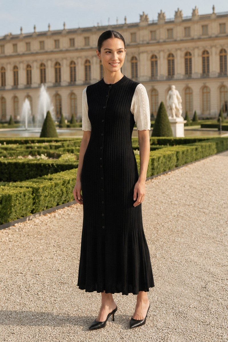 A woman stands outdoors in front of a grand palace and manicured gardens, wearing a sleeveless black pleated midi dress with short white sleeves and black pointed heels.